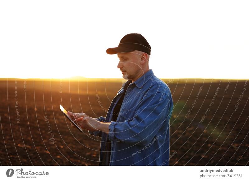 Intelligente Landtechnik und Landwirtschaft. Ein Landwirt benutzt ein digitales Tablet auf einem Feld mit gepflügtem Boden bei Sonnenuntergang. Überprüfung und Kontrolle der Bodenqualität, der Bereitschaft zur Aussaat von Pflanzen und zum Anbau von Gemüse