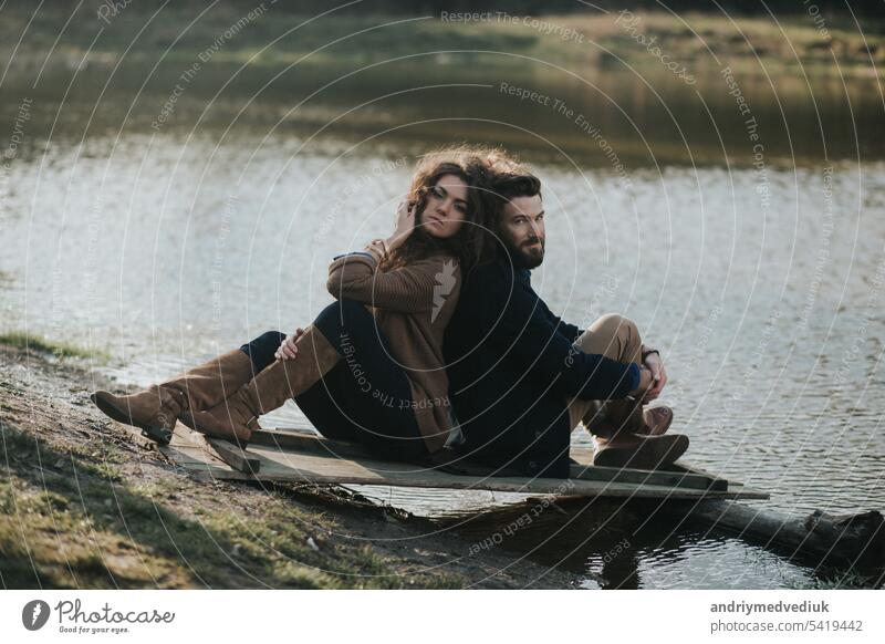 zwei kaukasische Liebhaber sitzen auf dem Pier am See. Junges Paar umarmt sich an einem Herbsttag im Freien. Ein bärtiger Mann und eine lockige Frau in Liebe. Valentinstag. Konzept der Liebe.