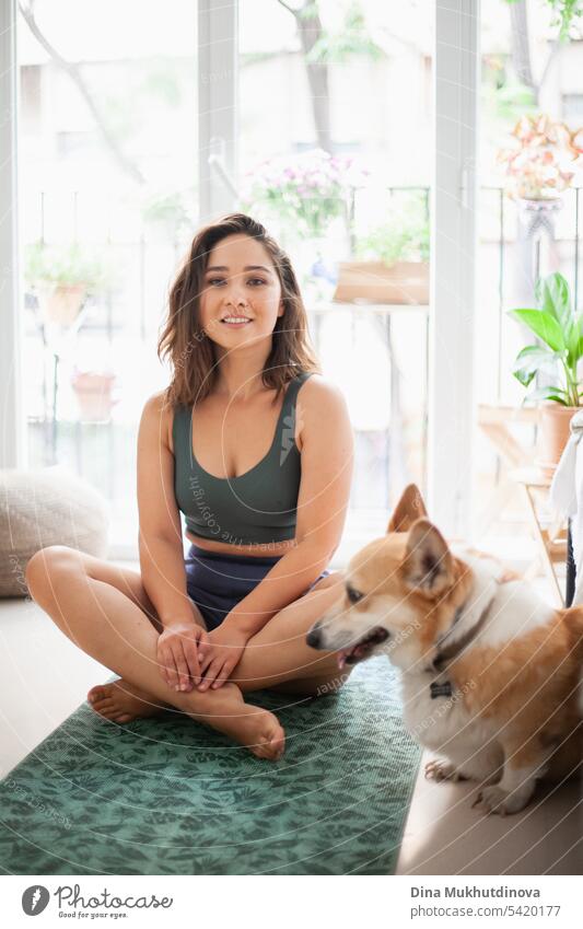Junge Frau mit Corgi Pembroke Hund macht Yoga auf grüner Matte zu Hause. Praktiziert Sport, Wellness und Gesundheit. Indoor Yoga Online-Kurs. Achtsamkeit und Meditation, aktiver Lebensstil.