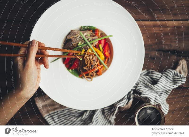 Person, die asiatische Nudeln mit Stäbchen isst Lebensmittel Speise Essstäbchen essen Hand Fleisch Gemüse Mahlzeit lecker hölzern weiß Teller Abendessen