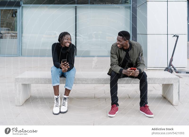 Junges schwarzes Paar trifft sich auf einer Straße in der Stadt Sitzung Großstadt Bank Lächeln Bekanntschaft ethnisch Smartphone urban sitzen Mann Frau Süchtige