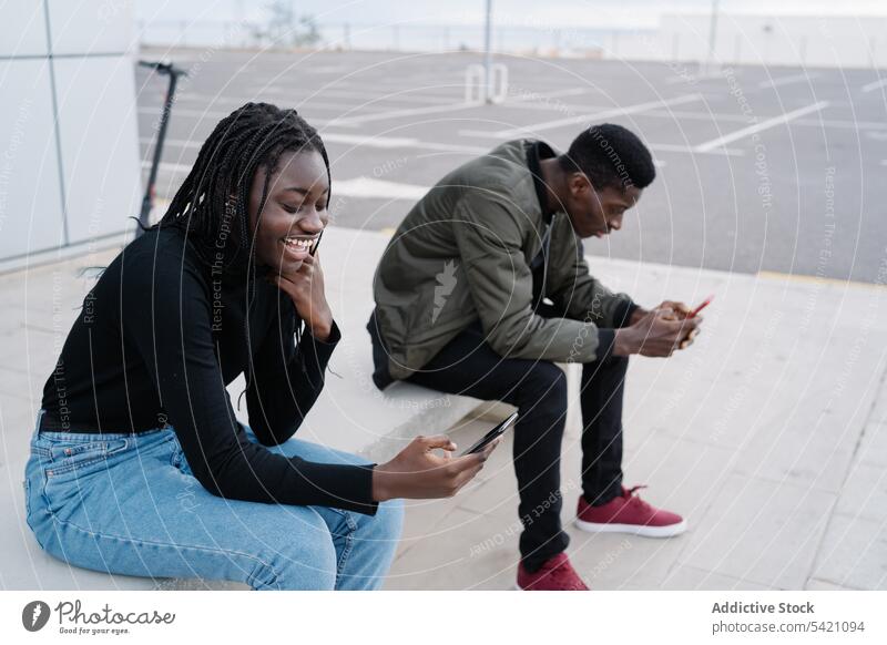Ethnisches Paar mit Smartphones auf einer Bank benutzend Straße sitzen Großstadt ethnisch urban Termin & Datum Mann Frau modern lässig schwarz Afroamerikaner