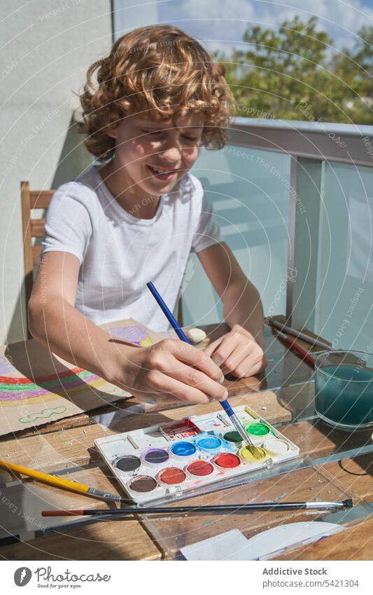 Fröhlicher Junge malt auf dem Balkon Farbe Wasserfarbe Tisch Lächeln Container Bürste Kunst kreativ Kind zeichnen Kindheit Werkzeug sitzen Glück Lifestyle
