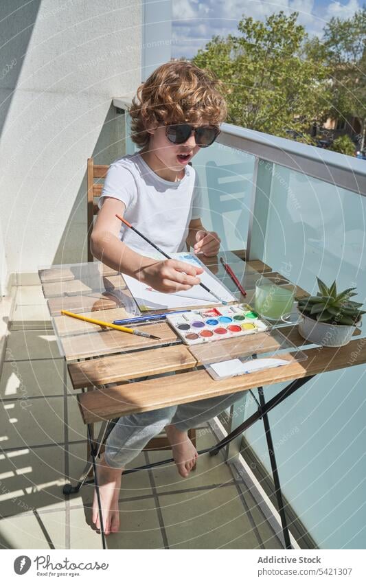 Fröhlicher Junge malt auf dem Balkon Farbe Wasserfarbe Tisch Lächeln Container Bürste Kunst kreativ Kind zeichnen Kindheit Werkzeug sitzen Glück Lifestyle