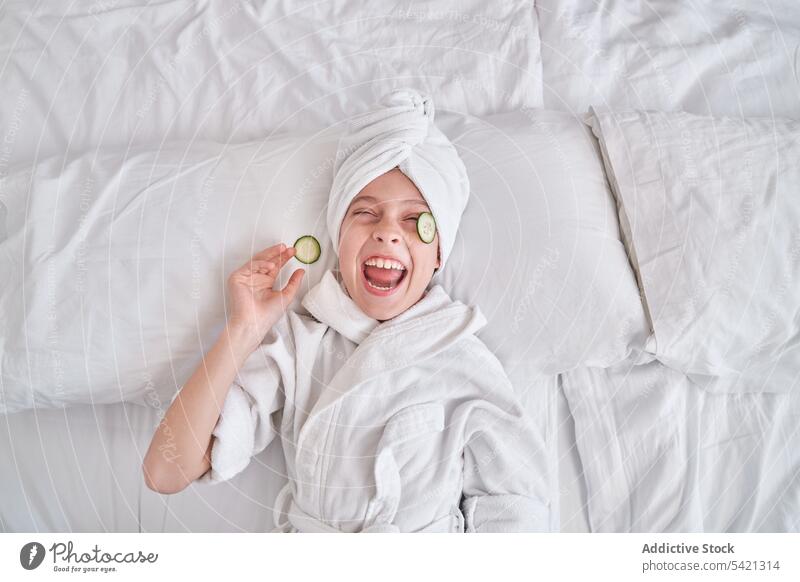 Fröhliches Kind mit Gurkenscheiben auf ja auf dem Bett liegend Salatgurke Mundschutz Schönheit Hautpflege Spaß positiv Lachen genießen Gesichtsbehandlung Witz