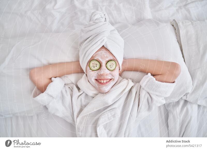 Verspieltes Kind mit Gesichtsmaske auf dem Bett liegend Mundschutz Gesichtsbehandlung Spaß haben Salatgurke Schönheit Hautpflege Grimasse spielerisch