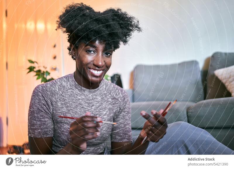 Fröhliche ethnische Frau malt zu Hause Farbe heimwärts positiv jung lässig Bürste Porträt Lächeln zeichnen Afroamerikaner schwarz Amateur Freizeit Hobby Glück