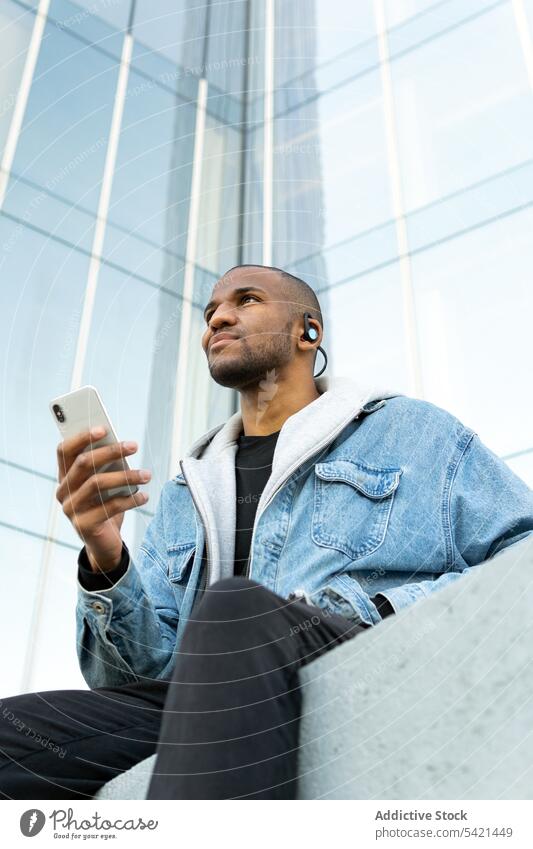 Schwarzer Mann mit Kopfhörern und Smartphone vor einem städtischen Gebäude zuhören Gesang Musik genießen Hand-in-Tasche Stadt benutzend Apparatur Gerät Klang