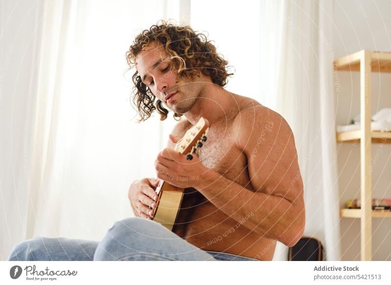 Mann ohne Hemd spielt Ukulele auf dem Bett zu Hause spielen Musik Musiker nackter Torso gutaussehend Hobby Instrument männlich sich[Akk] entspannen sitzen
