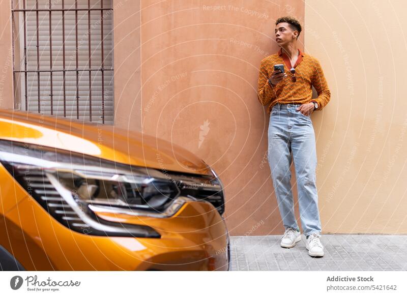 Ethnischer Mann mit Smartphone steht in der Nähe eines modernen Autos Stil Farbe PKW urban orange Automobil benutzend jung männlich ethnisch trendy Mobile