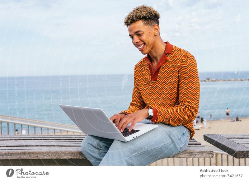 Gut gelaunter junger Mann mit Laptop am Strand benutzend Glück Schüler freiberuflich Stil MEER Tippen positiv ethnisch trendy Lächeln Afro-Look krause Haare