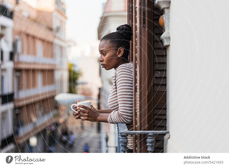 Schwarze Frau mit Kaffee, die vom Balkon aus auf die Stadt blickt reflektierend wehmütig Heißgetränk nachdenklich einsam Haus nachdenken allein Afroamerikaner