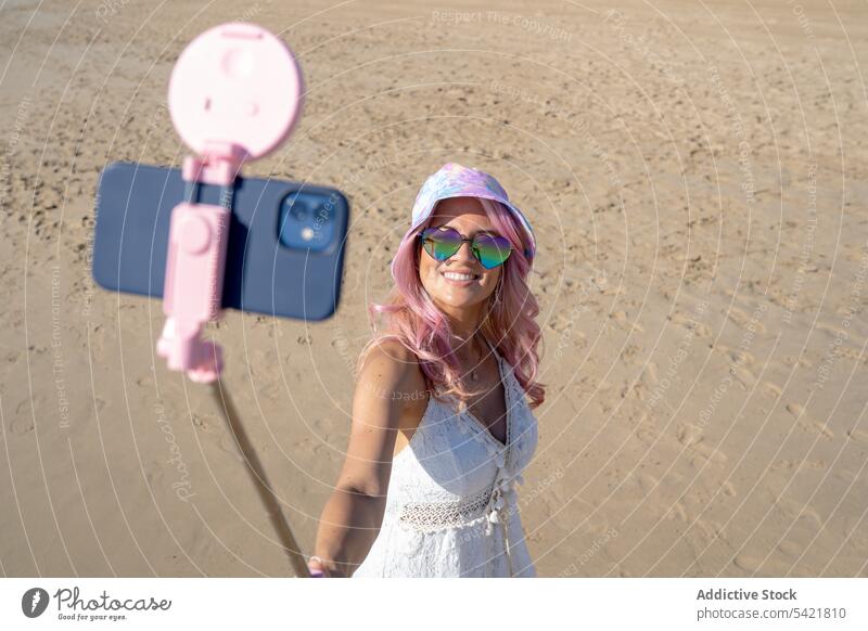 Lächelnde Frau mit rosa Haaren nimmt Selfie am Strand Sommer Selfie-Stick Smartphone Selbstportrait Meeresufer heiter Spaß haben fotografieren Apparatur Gerät