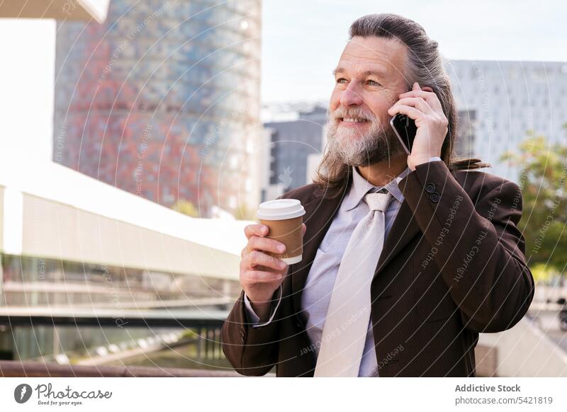 Fröhlicher bärtiger Geschäftsmann mit Kaffee im Gespräch mit Smartphone auf der Straße reden urban Lächeln Telefon Imbissbude modern Mann reif Lebensmitte
