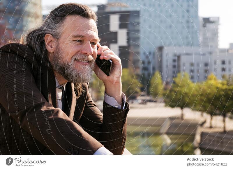 Glücklicher bärtiger Geschäftsmann, der in der Stadt mit seinem Smartphone spricht reden urban Lächeln Telefon Kommunizieren Vollbart Stil modern Mann reif