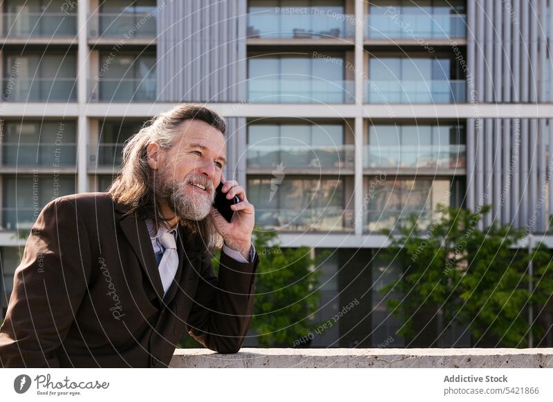 Glücklicher bärtiger Geschäftsmann, der in der Stadt mit seinem Smartphone spricht reden urban Lächeln Telefon Kommunizieren Vollbart Stil modern Mann reif