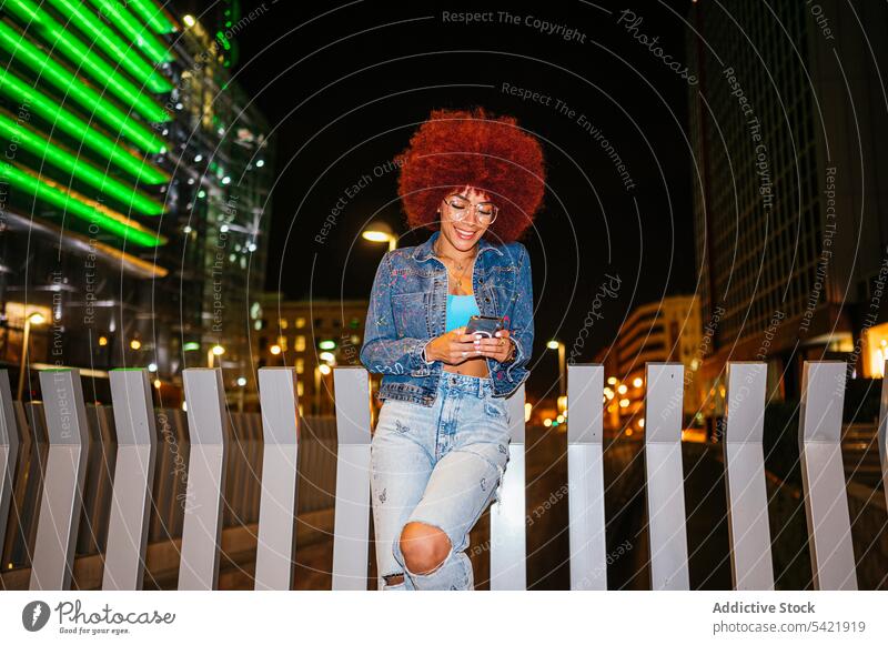 Stilvolle Frau, die auf einer dunklen Straße mit Gebäuden auf ihrem Smartphone surft Browsen online Afro-Look Abend Fechten Mode urban rendy Bekleidung Surfen