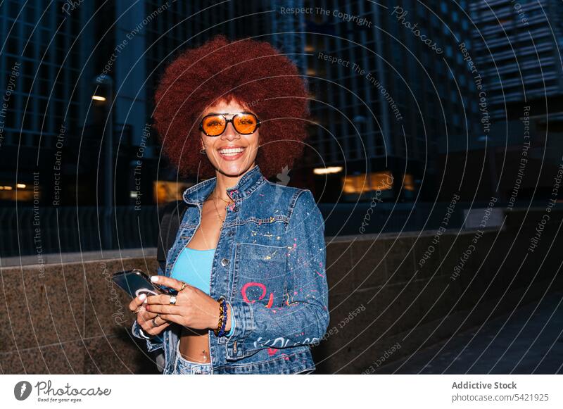 Stilvolle Frau, die auf der Straße ihr Smartphone durchsucht Browsen online Afro-Look Abend Gebäude urban Mode Haus trendy Bekleidung Surfen Textnachricht