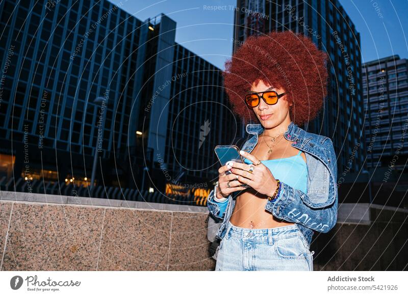 Stilvolle Frau, die auf der Straße ihr Smartphone durchsucht Browsen online Afro-Look Abend Gebäude urban Mode Haus trendy Bekleidung Surfen Textnachricht
