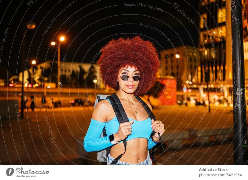 Stilvolle Frau mit Rucksack auf dunkler Straße Afro-Look Mode Bekleidung Outfit Gebäude Abend ernst selbstbewusst selbstsicher schlanke dunkel Dämmerung Haus