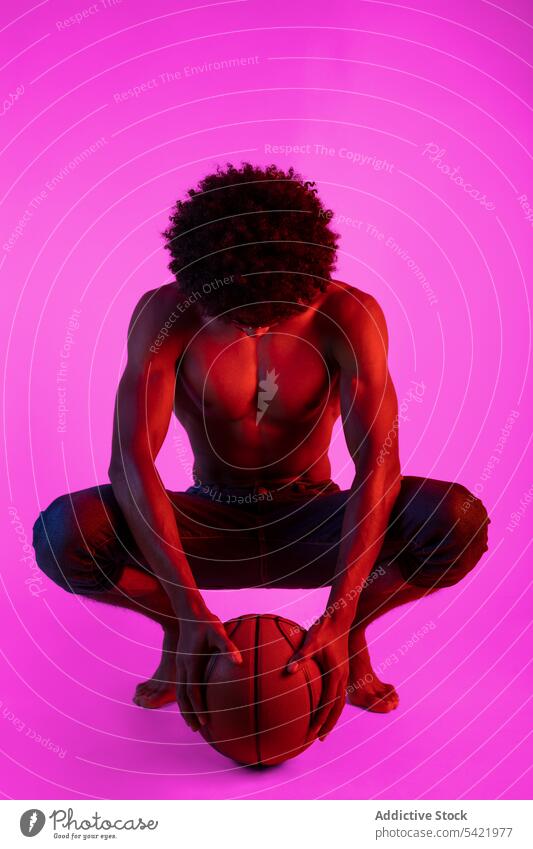Schwarzer Mann ohne Hemd mit Basketball sitzend nackter Torso muskulös Sportler Vorschein Spieler neonfarbig Kniebeuge männlich Afroamerikaner schwarz