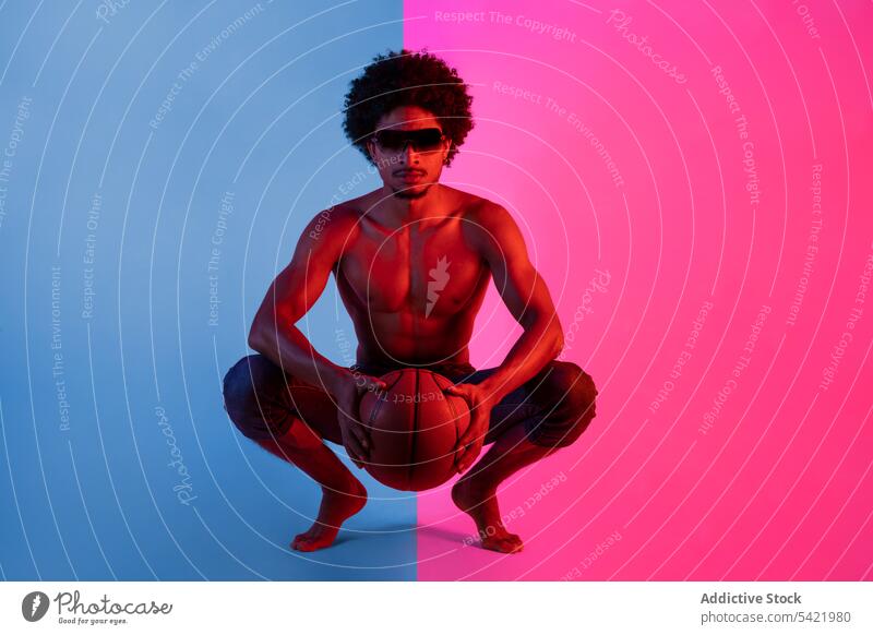 Schwarzer Mann ohne Hemd mit Basketball sitzend nackter Torso muskulös Sportler Vorschein Spieler neonfarbig Kniebeuge männlich Afroamerikaner schwarz