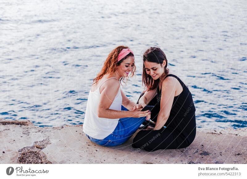 Zwei Freundinnen sitzen am Strand, benutzen ein Handy und lächeln. Technologie und Lifestyle Konzept Freunde lässig Telefon stylisch heiter Mobile positiv