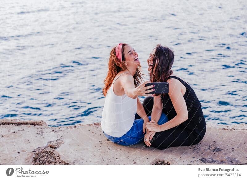 Zwei Freundinnen sitzen am Strand und benutzen ihr Handy, machen ein Selfie und lächeln. Technologie und Lifestyle Konzept Freunde lässig Telefon stylisch