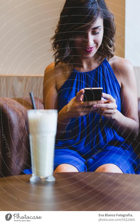 Junge schöne Frau genießt ein Smoothie-Getränk in einem Café und benutzt ein Mobiltelefon. Lässiges blaues Kleid. Modernes Leben in Innenräumen. selektiver Fokus