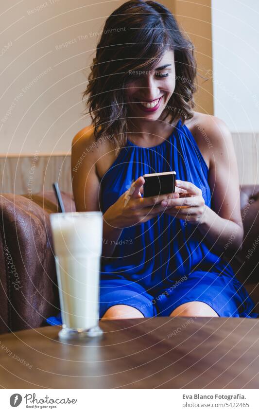 Junge schöne Frau genießt ein Smoothie-Getränk in einem Café und benutzt ein Mobiltelefon. Lässiges blaues Kleid. Modernes Leben in Innenräumen. selektiver Fokus