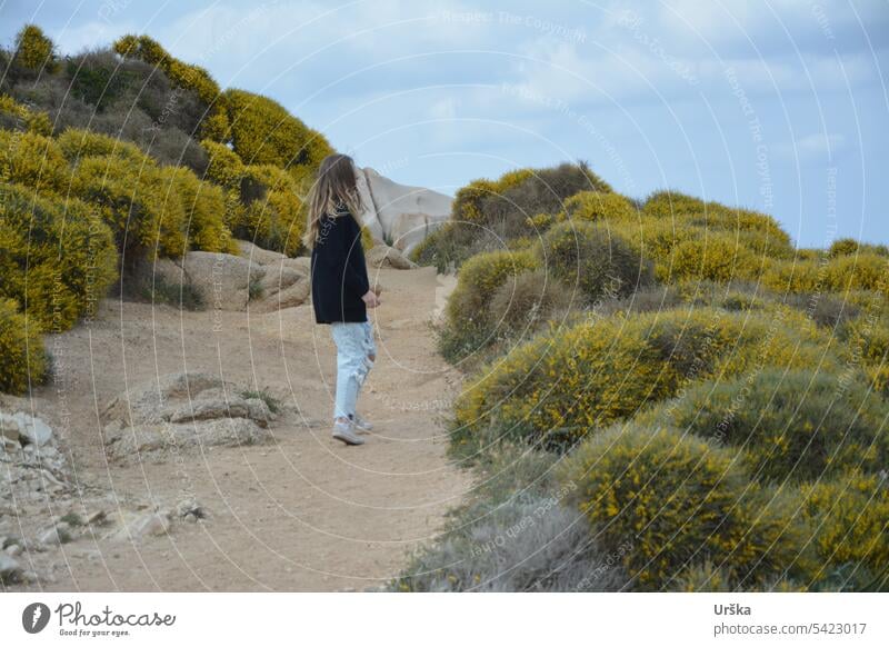 Ein Mädchen auf einem Weg zwischen Blumen in Sardinien Natur reisen lange Haare gelb Pflanzen Steine sardegna Sträucher Frühling Tageslicht