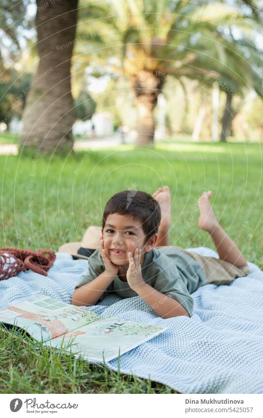 diverse Kind liest ein Buch im Park auf der blauen Decke auf grünem Gras, lächelnd und Blick in die Kamera. Fünf oder sechs Jahre alter Junge im Park im Sommer.