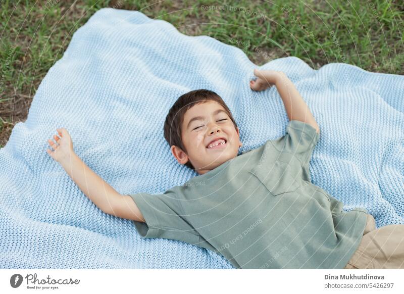 vielfältiges Kind im Park auf der blauen Decke liegend und lachend auf grünem Gras, lächelnd mit geschlossenen Augen. Fünf Jahre alter Junge im Park im Sommer, glücklich fühlen.