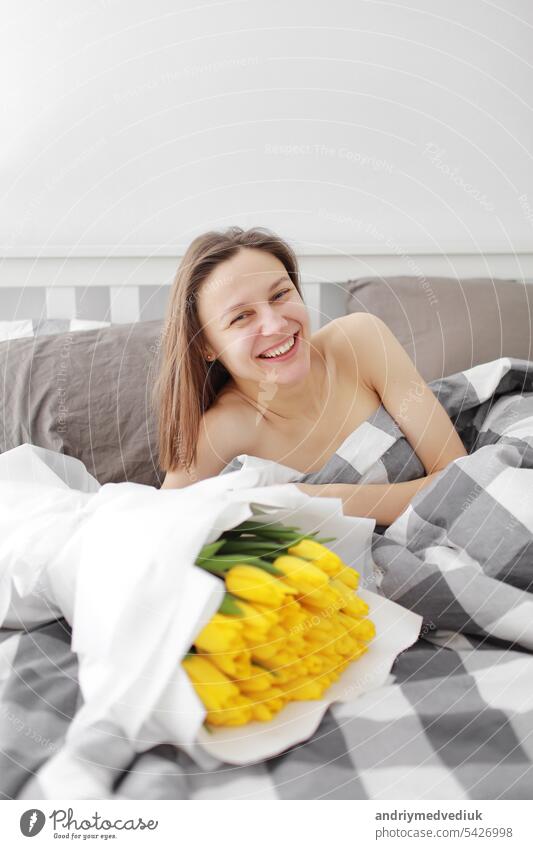 glückliches Mädchen mit Blumen gelbe Tulpen auf einem Bett liegend mit dem Genießen von Blumen und romantisches Geschenk in Valentinstag. Sie gerade aufgewacht und haben ihr einen Blumenstrauß präsentiert. Ein Geburtstag, Tag der Frau,
