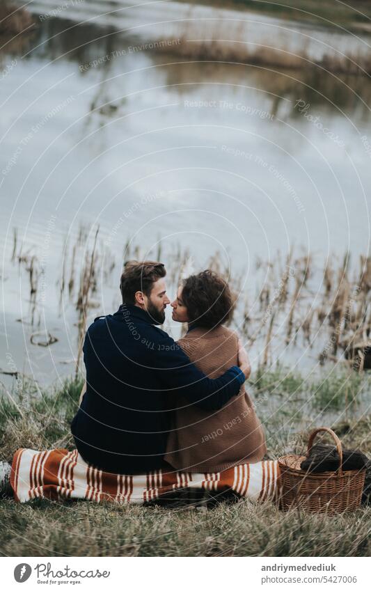 zwei kaukasische Liebhaber sitzen auf Decke am See. Junges Paar umarmt sich am Herbsttag im Freien. Ein bärtiger Mann und lockige Frau in der Liebe. Valentinstag. Konzept der Liebe und Familie. Rückansicht