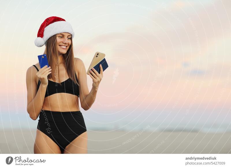 Glückliche junge Frau in Santa Hut und Badeanzug mit Smartphone, Kreditkarte und Reisepass ist die Buchung Online-Ticket für die Reise Weihnachten und Neujahr Urlaub am Strand. Rabatt Reisen Konzept.
