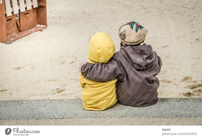 Zwei Kinder sitzen am Strand und passen aufeinander auf Brüder Verbundenheit Sand Sandstrand Regenjacke aufeinander aufpassen Freundschaft Außenaufnahme