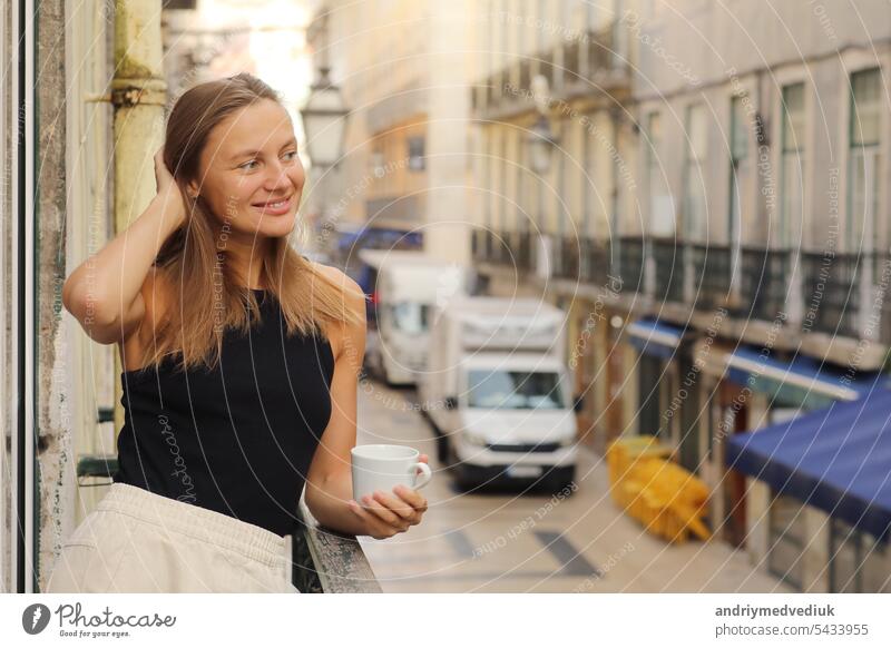 Glückliche junge schöne Frau hält Tasse Kaffee oder Tee, stehend auf dem Balkon mit Europa Stadt Straße am Morgen. Reife Frau genießt Urlaub im Resort. Sommer, Wochenende, Tourismus, Reisen