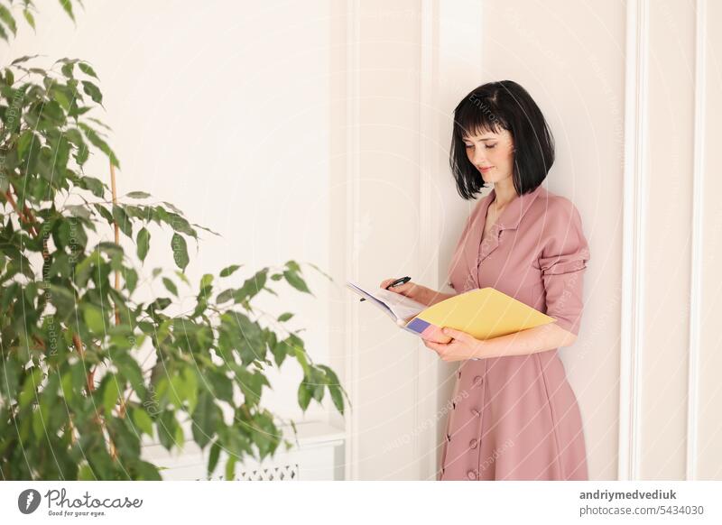 Junge brünette Frau in stilvollen weichen rosa Kleid ist in gelben Ordner von Papierkram mit mock up Bereich suchen und stehen im Büro mit Zimmerpflanzen. Bildung, Wirtschaft, Lifestyle-Konzept