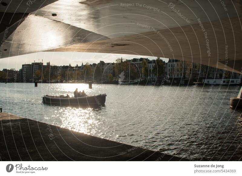Boot in der Sonne, fotografiert unter einer Brücke an den Amsterdamer Grachten. Die Sonne scheint, was zu einer Silhouette des Bootes führt. Auch die Brücke fängt die Sonne ein und glänzt. Das Bild hat eine idyllische, kühle und urbane Ausstrahlung.