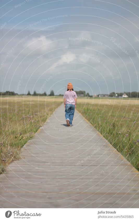 Mädchen läuft auf einem Steg Salzwiese Nordsee lässig Mütze hände taschen Jeanshose Sommer warm Holzsteg langer weg Blick nach vorn wolkiger himmel Gras grün