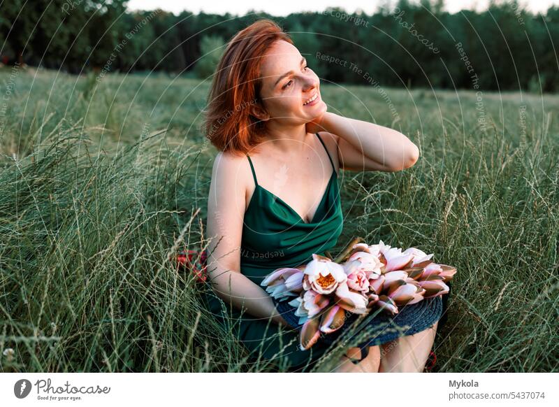 eine Frau, die sich im grünen Gras ausruht, ein langes grünes Kleid trägt und ein angenehmes Lächeln auf dem Gesicht hat. Sie hält Blumen in den Händen, genießt die Harmonie mit der Natur und erholt sich