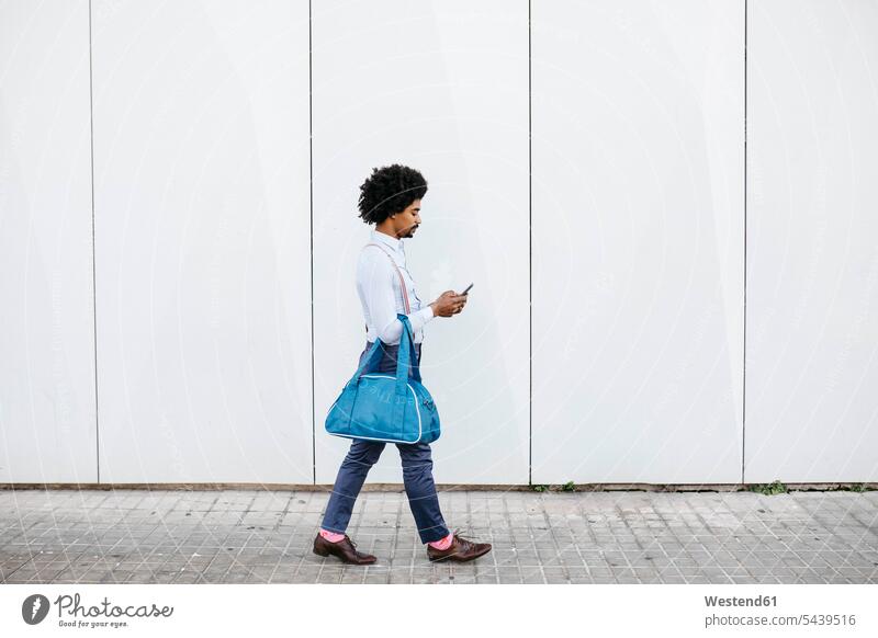 Mann mit Tasche geht vor einer weißen Wand, während er auf sein Handy schaut Männer männlich Taschen Mauer Mauern Smartphone iPhone Smartphones ansehen gehen