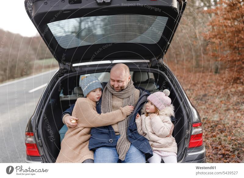 Lächelnder Vater mit zwei Kindern, die im Kofferraum eines Autos am Straßenrand sitzen Wagen PKWs Automobil Papas Väter Vati Vatis Papis Straßenränder