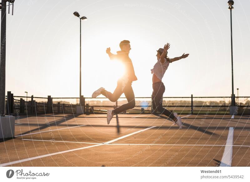 Unbeschwertes junges Paar springt bei Sonnenuntergang auf Parkdeck Sorglos Pärchen Paare Partnerschaft springen hüpfen Parkdecks Sonnenuntergänge Mensch