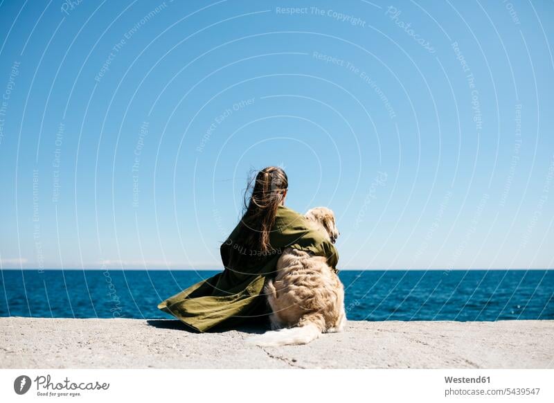 Rückenansicht einer Frau, die neben ihrem Labrador Retriever am Kai sitzt und auf den Horizont schaut Barcelona Entschleunigung Downshifting Einfaches Leben