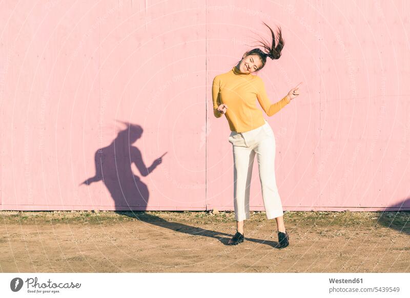 Glückliche junge Frau tanzt vor einer rosa Wand glücklich glücklich sein glücklichsein Wände Waende rosafarben weiblich Frauen Farbe Farbtöne Farbtoene Farbton