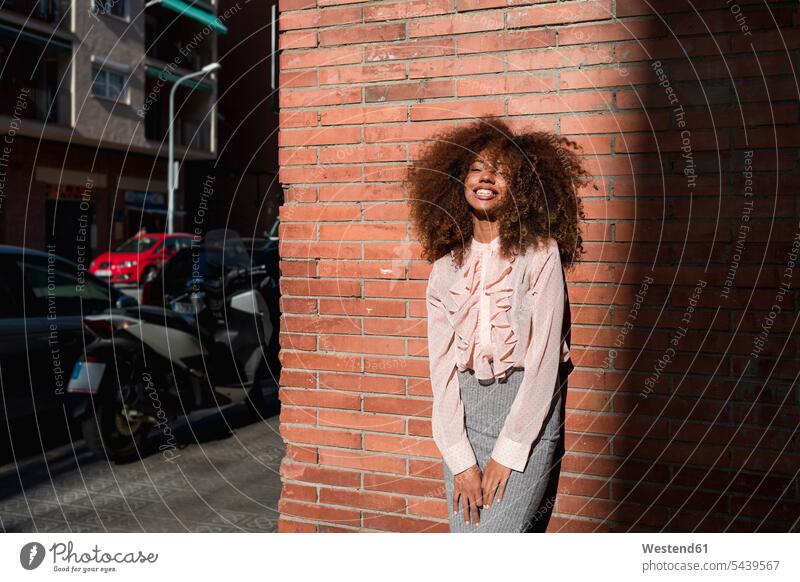 Porträt einer lächelnden jungen Frau mit Afro-Frisur, die an einer Ziegelmauer in der Stadt lehnt Portrait Porträts Portraits Afrolook Afros Afro-look