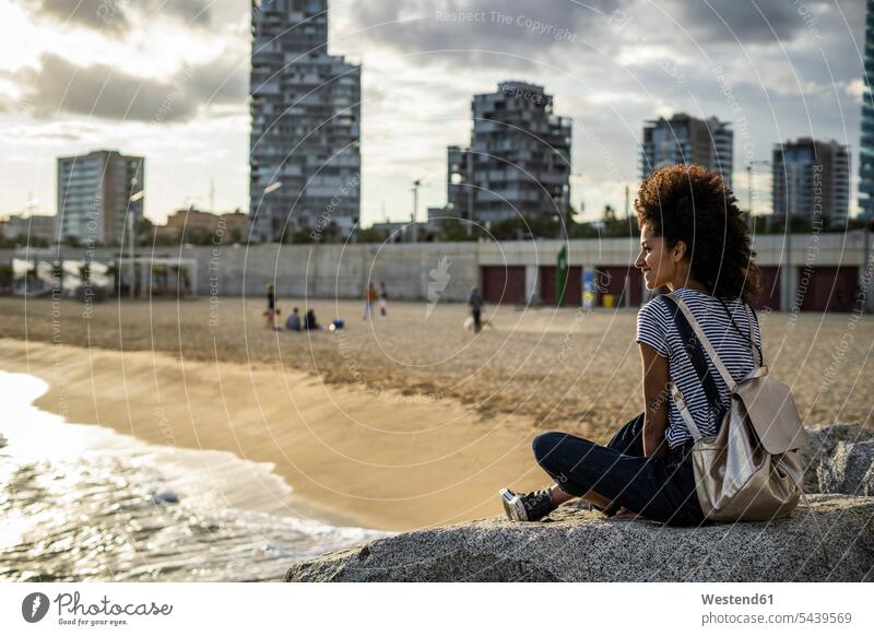 Junge Frau entspannt am Strand, sitzt auf Felsen, Rückansicht Afroamerikanisch Afro-Amerikanisch Afroamerikaner Afro-Amerikaner Ganzkörperansicht Ganzansicht