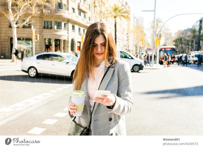 Spanien, Barcelona, junge Frau mit Kaffee, die am Straßenrand steht und auf ihr Handy schaut weiblich Frauen stehen stehend Smartphone iPhone Smartphones